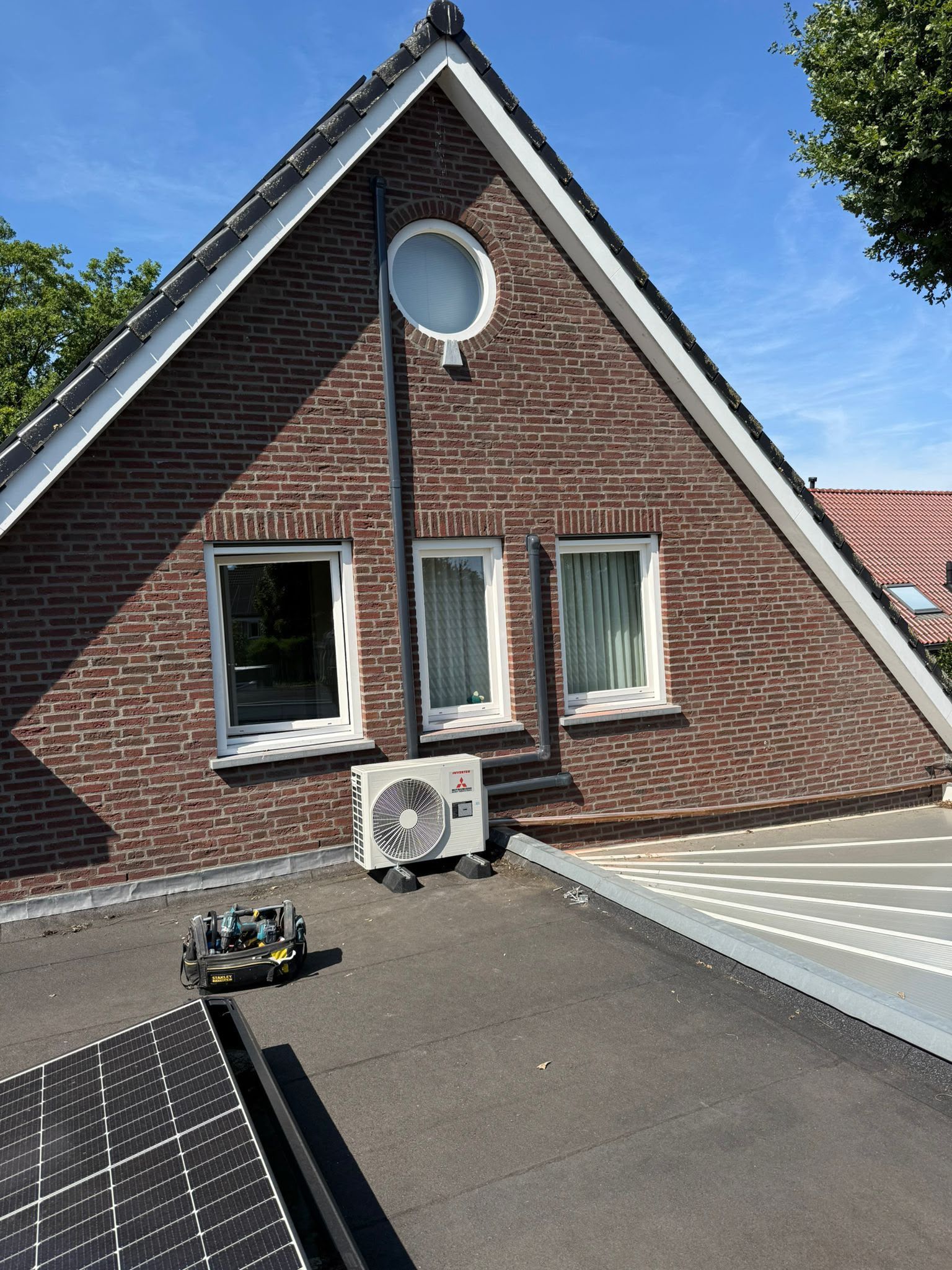 Witte buitenunit tegen een bakstenen puntgevel, op een plat dak naast zonnepanelen