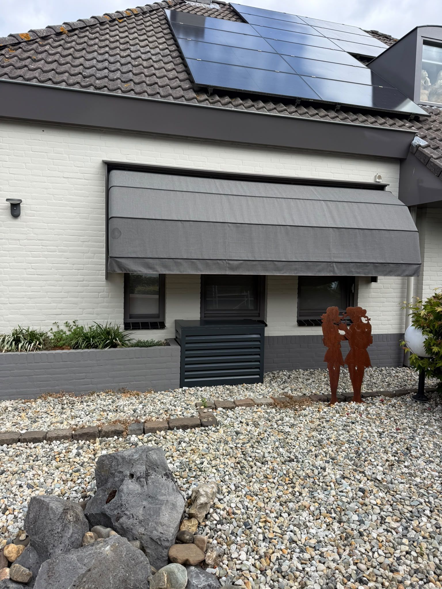Buitenunit in een zwarte omkasting tegen een witte stenen gevel, onder een grijze zonwering