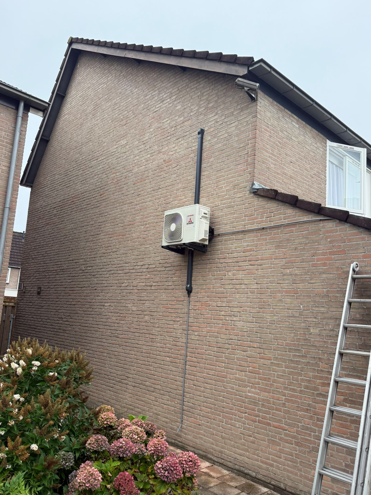 Witte buitenunit op muursteun tegen een bakstenen zijgevel, met leiding in een zwarte goot