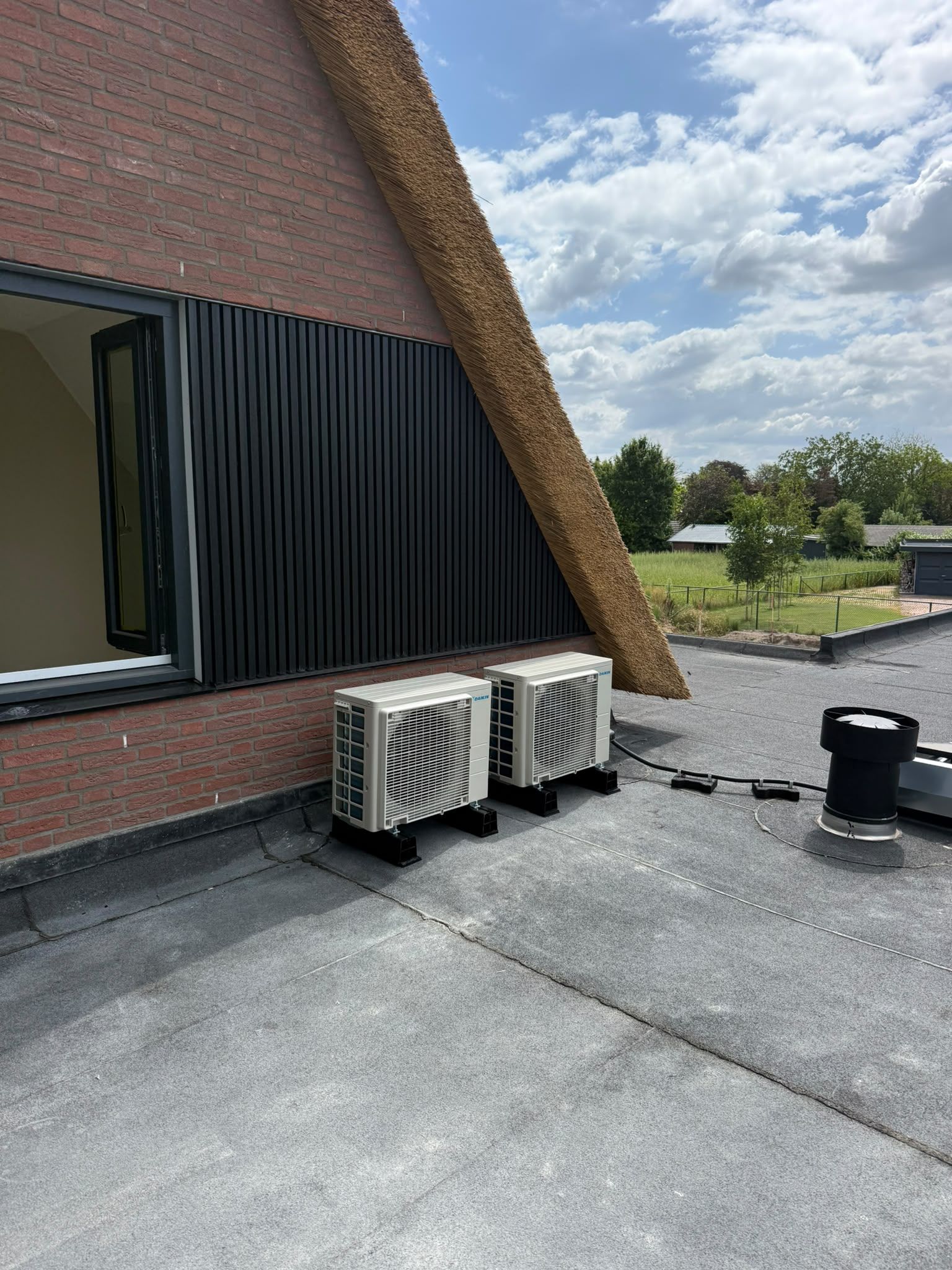 Twee witte buitenunits naast elkaar op een plat dak, langs een zwarte gevel onder een rieten dak